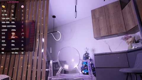Video of a modern, dimly lit kitchen with wooden cabinets, a clear acrylic chair, and a hanging wire light fixture, displaying a video game interface.