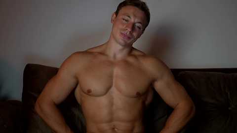Video of a shirtless, muscular, fair-skinned man with short brown hair, leaning back on a brown leather couch, eyes closed, against a plain white wall.