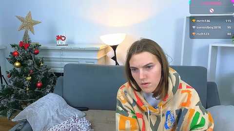 Video of a woman in a colorful, abstract-patterned hoodie, sitting on a gray couch, with a decorated Christmas tree in the background.