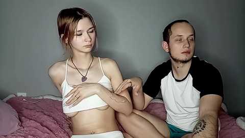 Media: Video of a young Caucasian woman with light skin and auburn hair, wearing a white bikini, sitting on a bed with a man in a black and white shirt, both looking sad.