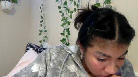 Video of a young woman with medium skin tone, dark hair in a messy bun, wearing a silver jacket, and a septum ring. Background features green vines and a white wall.