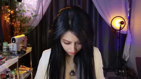 Video of a young Asian woman with long black hair, wearing a beige cardigan, focused on sewing. Background features dark curtains, a sewing machine, and a vintage lamp.