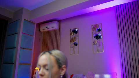 A video of a modern living room with a woman's blurred face in the foreground. The background features light purple and white walls, a vertical air conditioner, and decorative floral wall art.