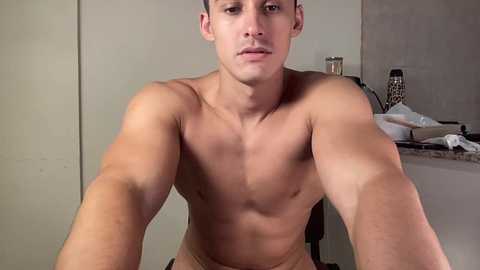 Video of a shirtless young man with short dark hair and a muscular build, sitting in a modern kitchen with beige walls and granite countertops.