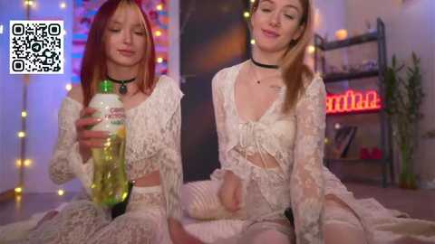 Video of two young women with fair skin, one with red hair, the other with blonde, both wearing lace lingerie, drinking green beer, in a dimly lit, cozy room with string lights and a neon sign.