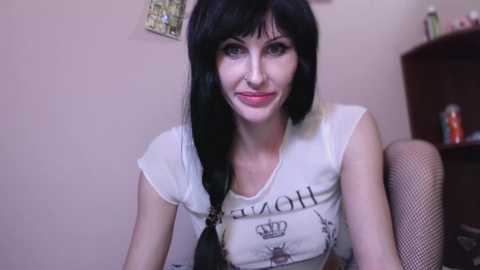 Media: Video of a slender, pale-skinned woman with black hair and green eyes, wearing a white T-shirt with a crown design and a black bra, seated indoors.