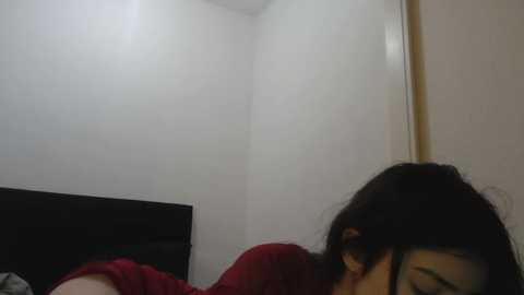 A video of a woman with dark hair and fair skin lying on a bed in a dimly lit, white-walled room, wearing a red shirt, with a visible black headboard.