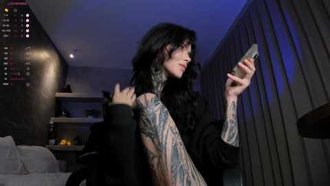 Video of a tattooed woman with long black hair, wearing a black dress, looking at her phone, in a dimly lit, modern room with gray walls and a TV screen showing game stats.