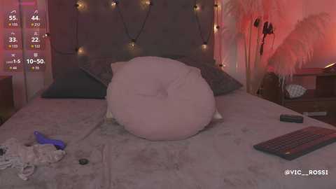 Video of a messy bedroom with a large pink pillow on a bed, a keyboard on the floor, and a thermometer on a nightstand. Warm ambient lighting and a wooden headboard.