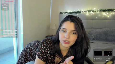 Video of a young woman with long dark hair, wearing a black floral dress, leaning over a bed in a modern, softly lit bedroom.