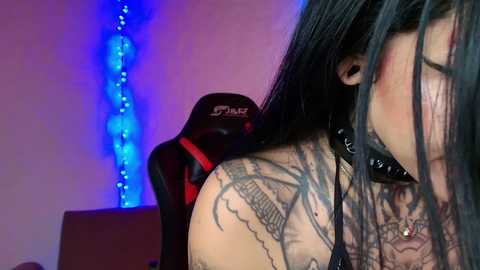 Video of a woman with long black hair, wearing a black collar, leaning against a red gaming chair with a blue LED strip background. Her tattooed shoulder is visible, and she has a black choker around her neck.