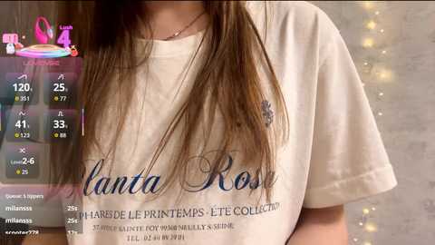 Video of a woman with long brown hair wearing a white T-shirt with \"Kantata Rose\" text. Background features a soft, blurred, beige wall with fairy lights.