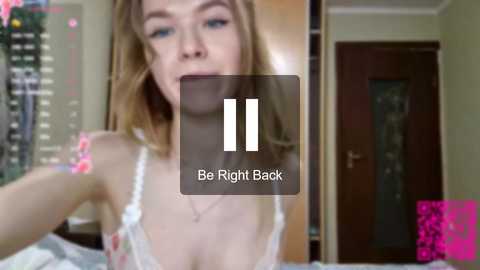 Video of a young woman with long blonde hair, wearing a white lace bra, in a room with a pink wall, a TV, and a door.