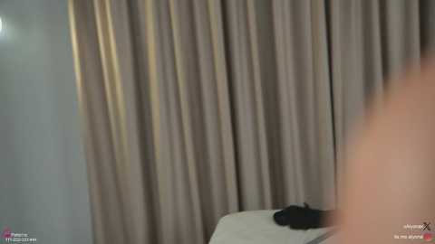 A video of a dimly lit bedroom with beige curtains, a white bed, and a partially visible person lying on their stomach, wearing black clothing.