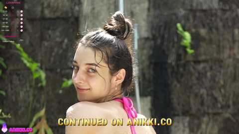 Video of a young woman with wet, dark hair tied up in a bun, smiling, wearing a pink bikini top, standing outdoors in front of a stone wall with green vines, with \"Continued on Anikoi.co\" text overlay.