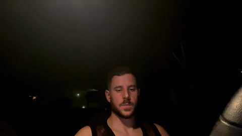 Video of a muscular, fair-skinned man with short hair and a beard, wearing a black tank top, standing in a dimly lit room with a spotlight on his face.