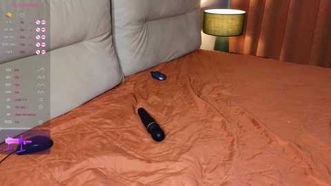 A video of a modern bedroom with a beige leather headboard, an orange bedspread, and a black vibrator on the bed. A calendar and a purple box are visible on the left.