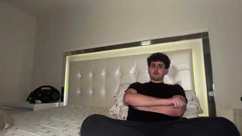 Video of a young man with short dark hair, wearing a black T-shirt, sitting on a bed in a modern bedroom with white bedding, a tufted headboard, and a black fan.