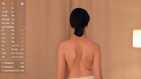 Video of a topless woman with dark hair tied back, viewed from behind, standing in a spa-like room with beige walls and a lit lamp.
