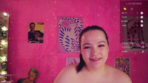 Video of a smiling, topless, fair-skinned woman with dark hair, in a pink room adorned with posters and a green plant.