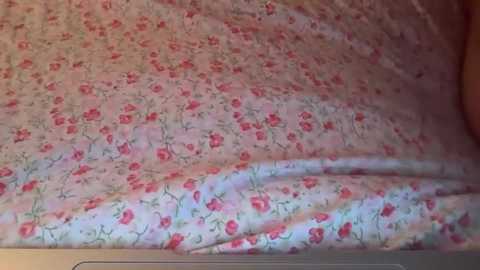 Video of a bed with a light pink floral sheet, featuring red roses and green leaves, slightly wrinkled and pulled down at the corners, showing a beige pillow peeking from the right side.
