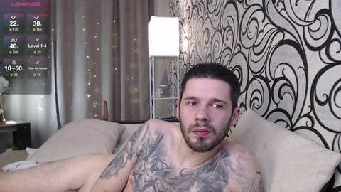 A video of a shirtless, tattooed man with a beard, lounging on a beige couch in a room with black and white patterned wallpaper and a lamp.