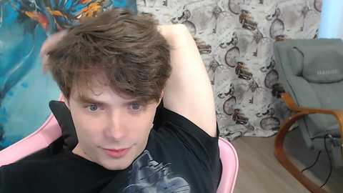 A video of a young, pale-skinned man with short brown hair, wearing a black T-shirt, sitting in a pink chair, with a relaxed pose, in a living room with patterned curtains and a gray couch.