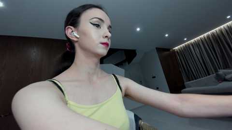 Video of a slim, light-skinned woman with dark hair, wearing a yellow tank top and black earbuds, standing in a modern, dimly lit room with brown walls and a sleek, gray couch.