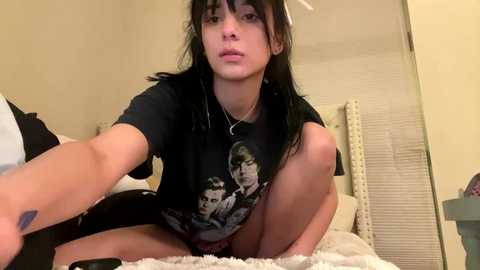 Video of a young woman with medium skin tone and black hair, sitting on a bed, wearing a black t-shirt with a graphic, white shorts, and a necklace. Background includes a white bedspread and beige walls.