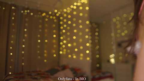 A video of a softly lit bedroom with warm yellow fairy lights, a bed with a colorful quilt, and a person's arm in the foreground.