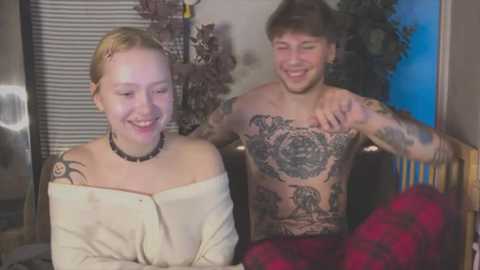 Media: Video of a smiling, tattooed young woman with blonde hair, wearing a cream top, and a man with tattoos, shirtless, embracing her in a cozy room with plants.