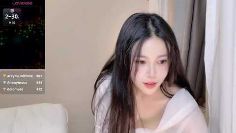 A video of an Asian woman with long black hair, fair skin, and a slender figure, wearing a white top, captured indoors against a beige background.