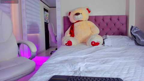 Video of a plush teddy bear with red ribbon and paw pads, sitting on a white bed with a tufted pink headboard, in a modern bedroom with white walls and purple lighting.