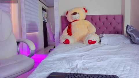Video of a modern bedroom with a plush teddy bear, a tufted pink headboard, white bedding, and a white chair with purple LED lights.