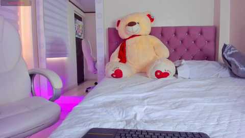 Video of a plush teddy bear with a red bowtie sitting on a bed with a pink tufted headboard, surrounded by white bedding and a purple glow.