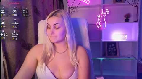 Video of a fair-skinned, blonde woman with medium-sized breasts, wearing a low-cut white top, sitting in a chair in a dimly lit room with neon lights and shelves.
