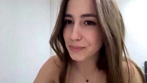 Media: A video of a young woman with long, straight, light brown hair, fair skin, and a neutral expression. She is topless, with a subtle, natural makeup look. The background is a plain, light-colored wall.