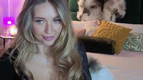 Video of a smiling, fair-skinned woman with long, wavy blonde hair, wearing a black fur-trimmed coat, in a cozy bedroom with a white bed, yellow pillows, and a lit lamp.