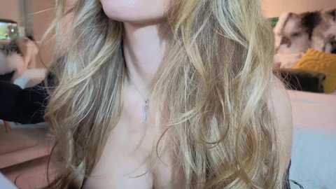 Video of a young woman with long, wavy, blonde hair, fair skin, and a slender build. She is wearing a necklace and is topless, with a blurred background featuring a bedroom with a bed and a person in a yellow sweater.