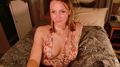 A video of a woman with light skin and shoulder-length brown hair, wearing a low-cut floral halter dress, sitting on a bed with a gray patterned blanket. The room has a wooden headboard and a nightstand with a lamp.