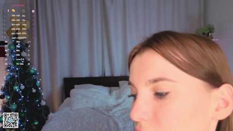 Video of a young woman with straight, light brown hair, eyes closed, in a dimly lit bedroom with a Christmas tree adorned with blue and silver ornaments and white curtains.