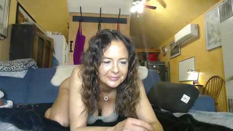 Video of a smiling, topless woman with curly brown hair, lying on a blue couch in a cluttered, cozy living room with yellow walls, framed art, and a ceiling fan.