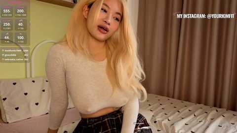 Video of a blonde, light-skinned woman with long hair, wearing a white long-sleeve crop top and a plaid skirt, sitting on a bed with a heart-patterned duvet in a cozy, beige-toned bedroom.
