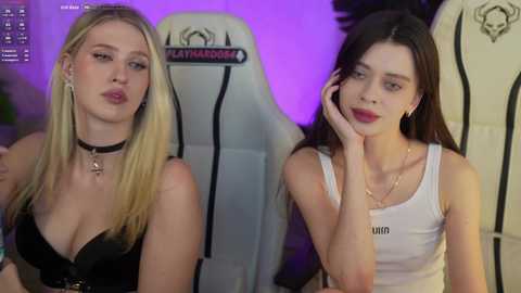Video of two young women seated on white gaming chairs; one blonde with a black choker, black top, and lips, the other brunette with a white tank top and red lips, against a purple-lit background.