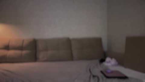 A blurred video of a minimalist bedroom with beige walls, a large beige headboard, a bed with white sheets, a side table with a book, and a soft, warm light.