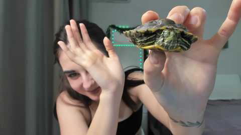 Video of a young woman with long brown hair, fair skin, and a small tattoo on her wrist, smiling while holding a green and brown snake close to her face.
