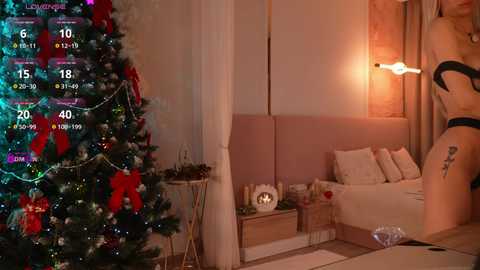 Video of a cozy, warmly lit bedroom with a Christmas tree adorned with red and white ornaments, a woman in a black bra and underwear, and a digital display showing a calendar.