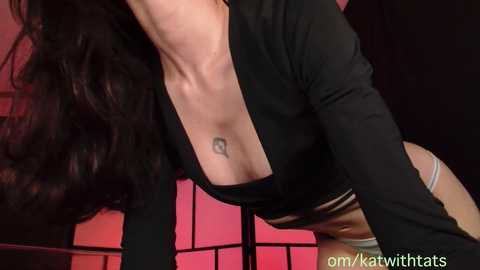 Video of a woman with long, dark hair, wearing a black, low-cut top, and a tattoo on her collarbone, against a red-lit background.