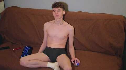 Video of a thin, young, shirtless white male with short brown hair, wearing black briefs and white socks, sitting on a brown couch with a blue mouse and red remote control beside him.