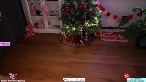 A video of a cozy living room with a decorated Christmas tree, red ornaments, and string lights, placed on a wooden floor. The background features a white bookshelf and a pink heart banner.